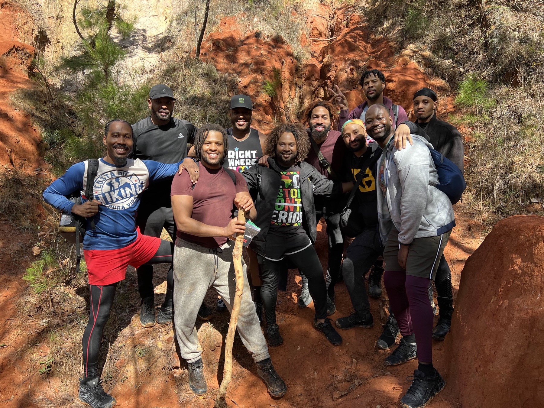 Black men smile together inside a sunlit red-clay canyon lined with pine trees.