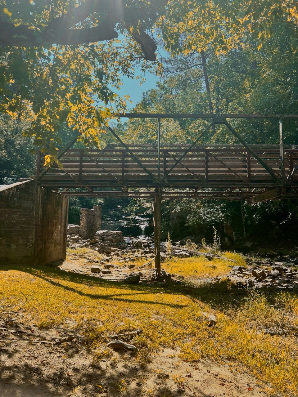 A metal footbridge spans a rocky creek beneath green and golden leaves.