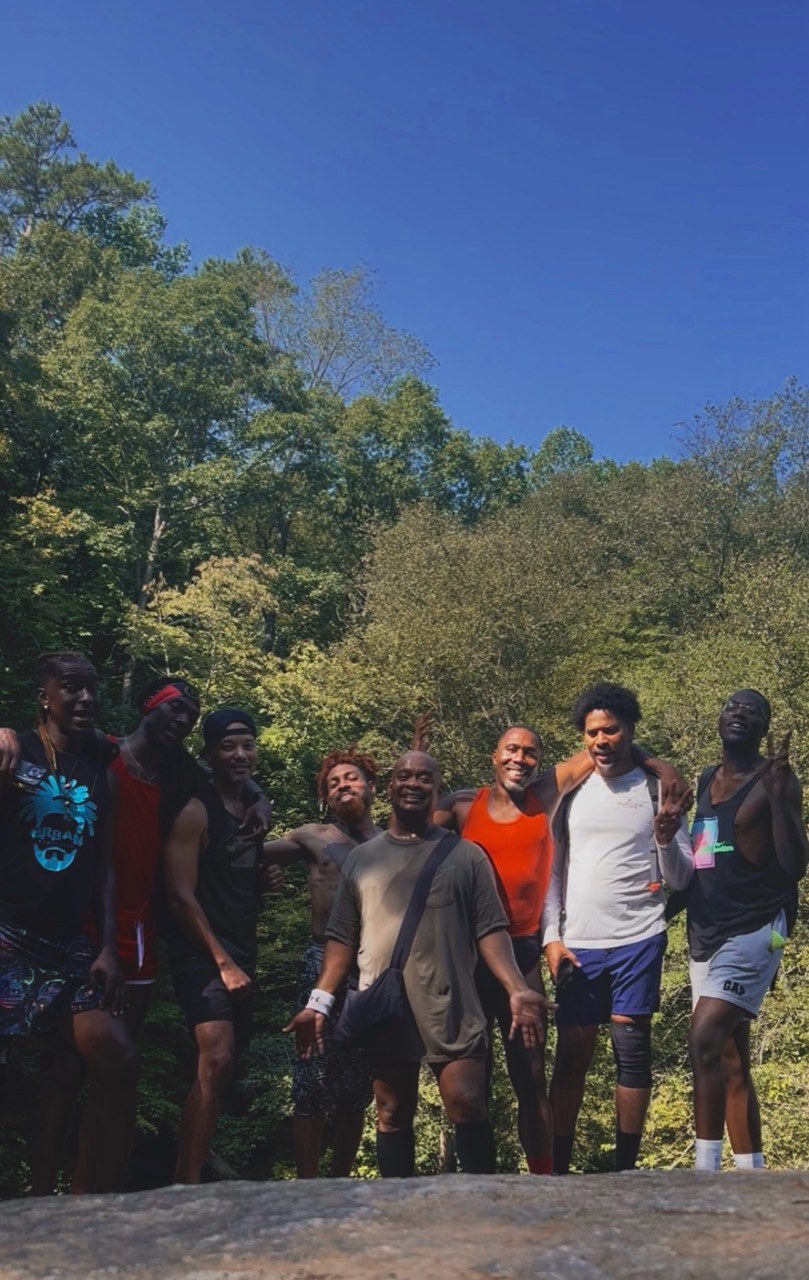 Eight hikers smile together on a rock outcrop with sunlit trees behind them.