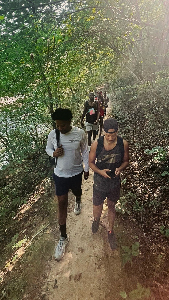 Hikers follow a narrow dirt trail beside the creek under a leafy canopy.