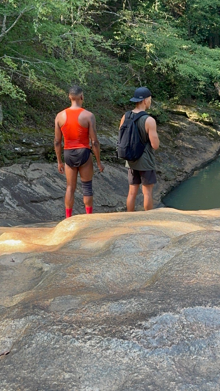 Two hikers stand on sloping rock beside a green creek pool bordered by trees.
