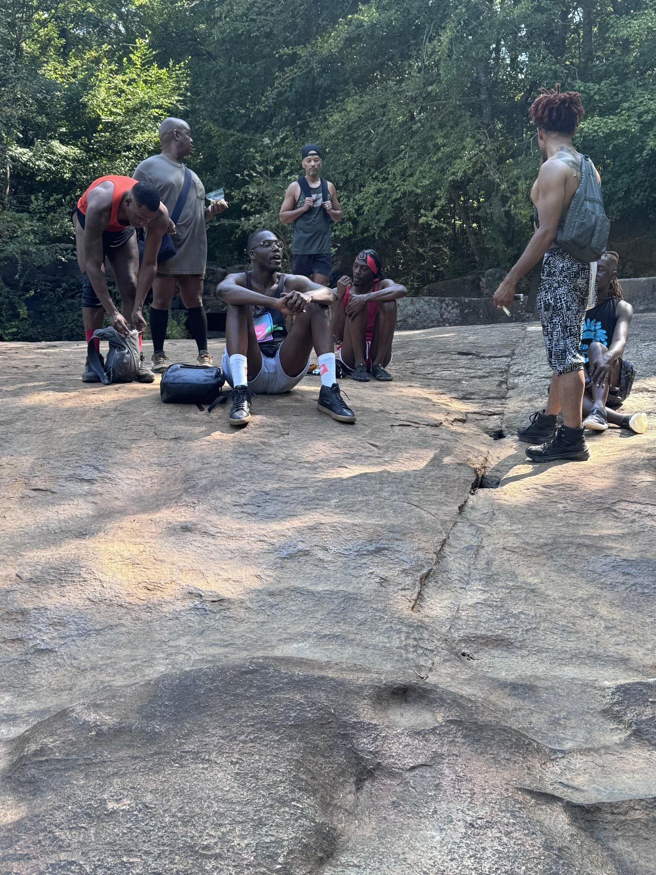 Hikers sit and stand together during a break on a broad rock outcrop surrounded by forest.