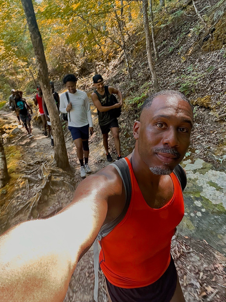 A hiker in an orange tank top takes a selfie with the group walking the wooded trail behind him.