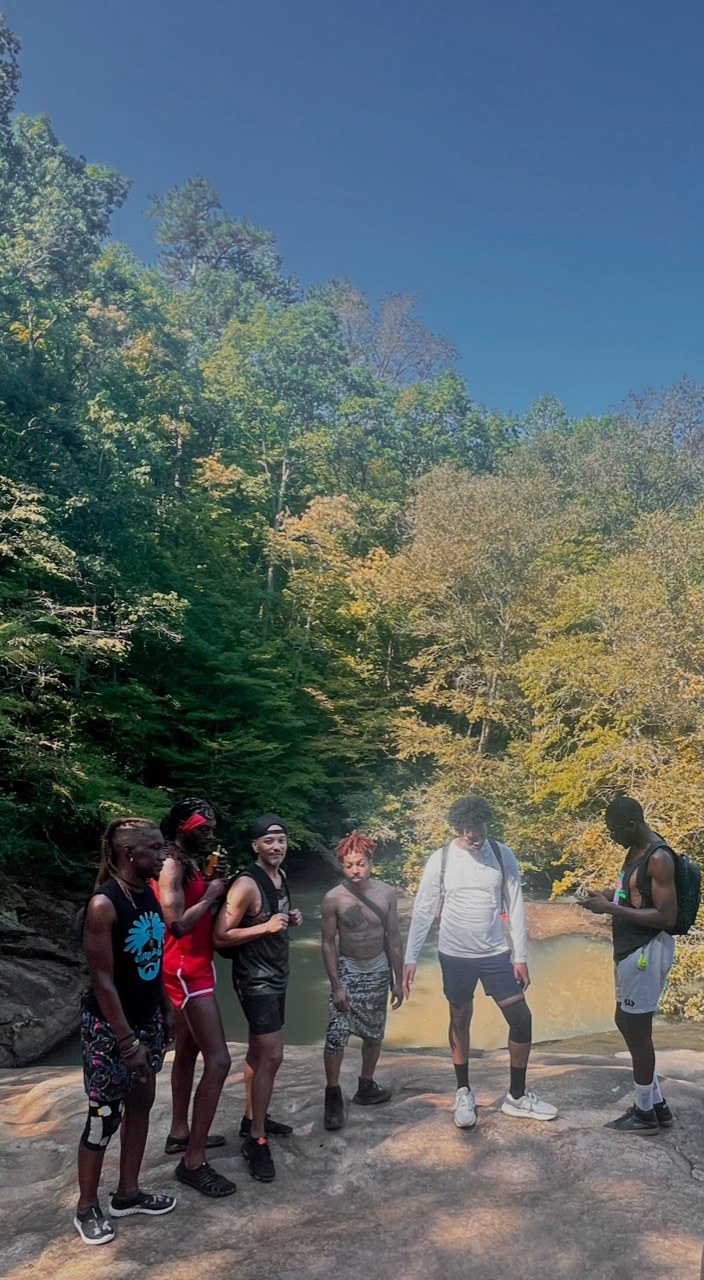 Six hikers gather on sunlit rock beside the water beneath a clear blue sky.