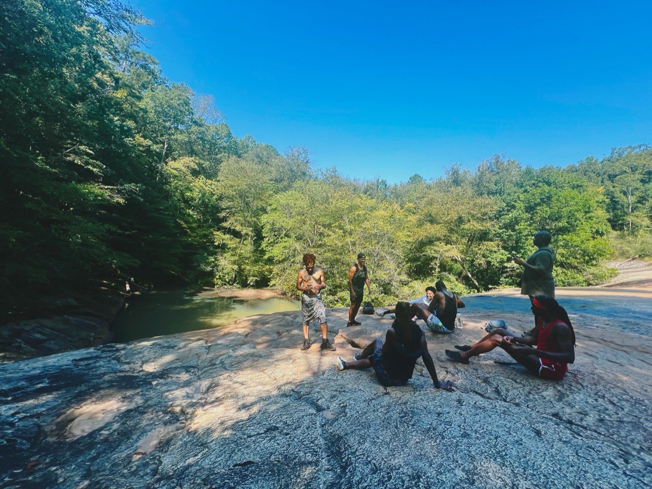 The group relaxes across a wide rock outcrop beside a forest creek pool.