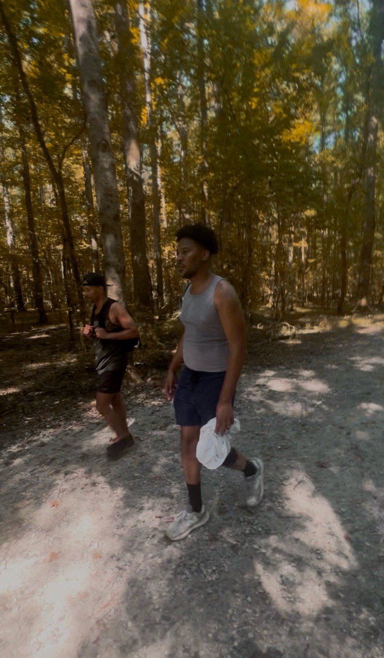 Two hikers walk a wide dirt path through sun-dappled woods.