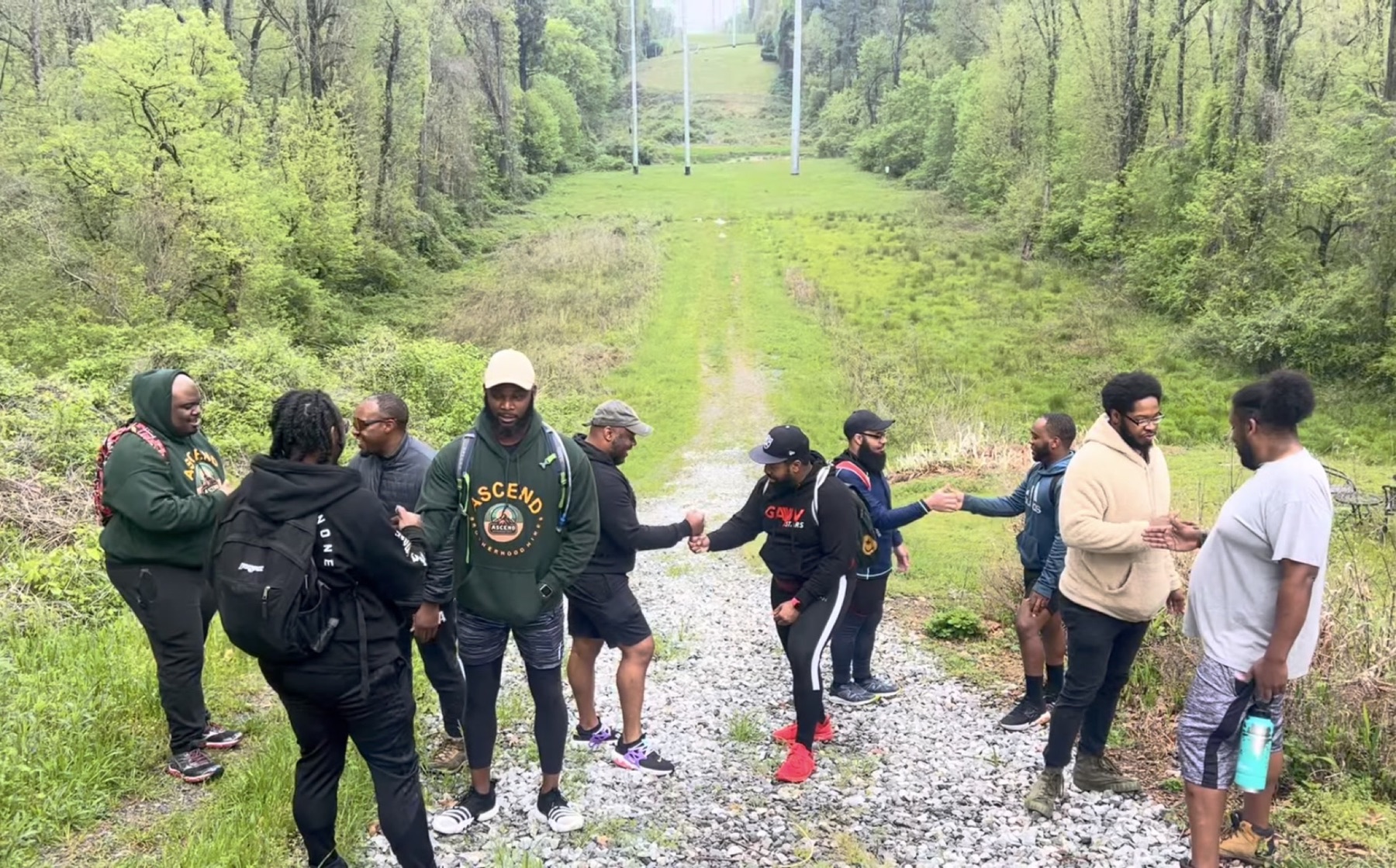 Black men greet one another on a gravel path beneath a green utility corridor.