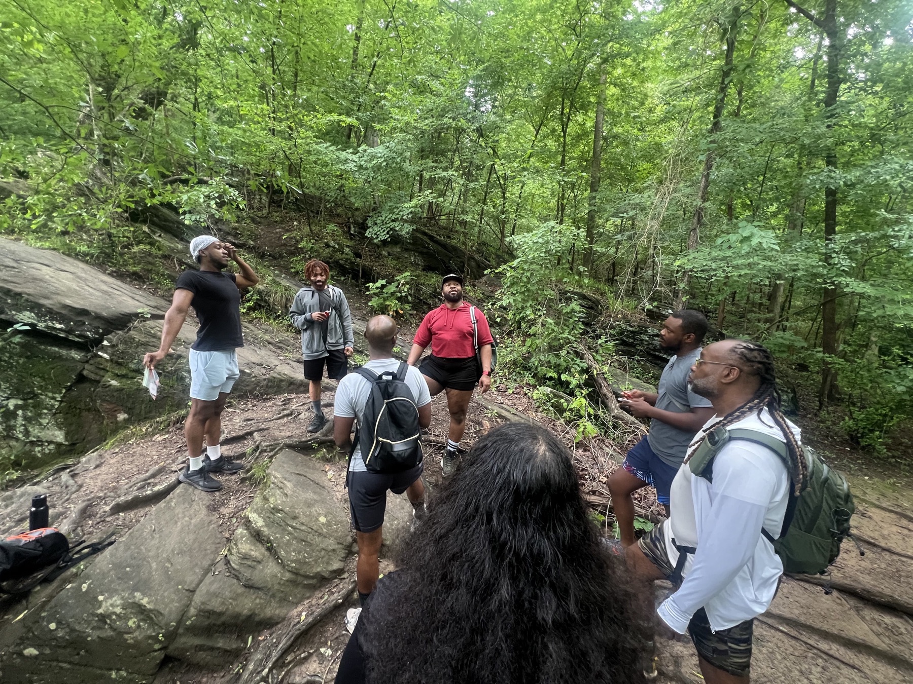 Black men pause together on a rocky forest slope surrounded by dense summer foliage.
