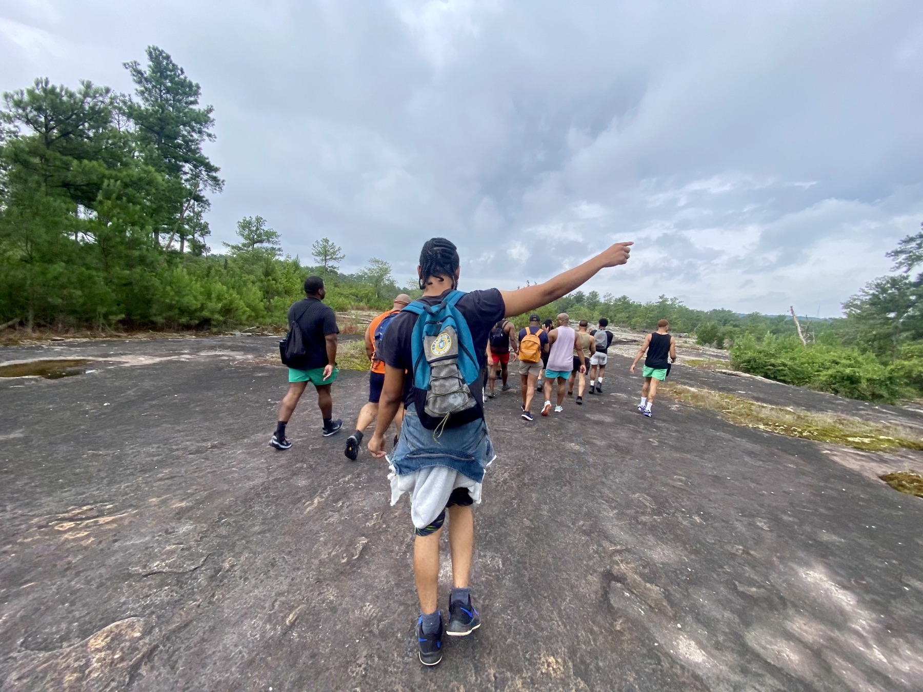 A Black hiker raises his arm while leading a group across a broad granite outcrop under cloudy skies.