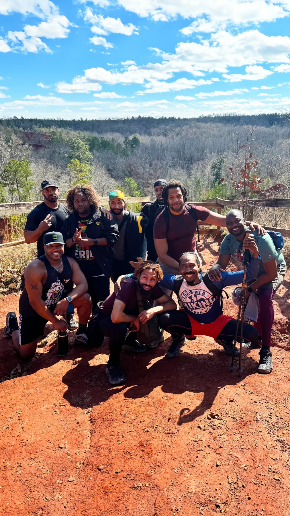 A large group of Black men pose together on a sunlit red-clay overlook with forested hills behind them.