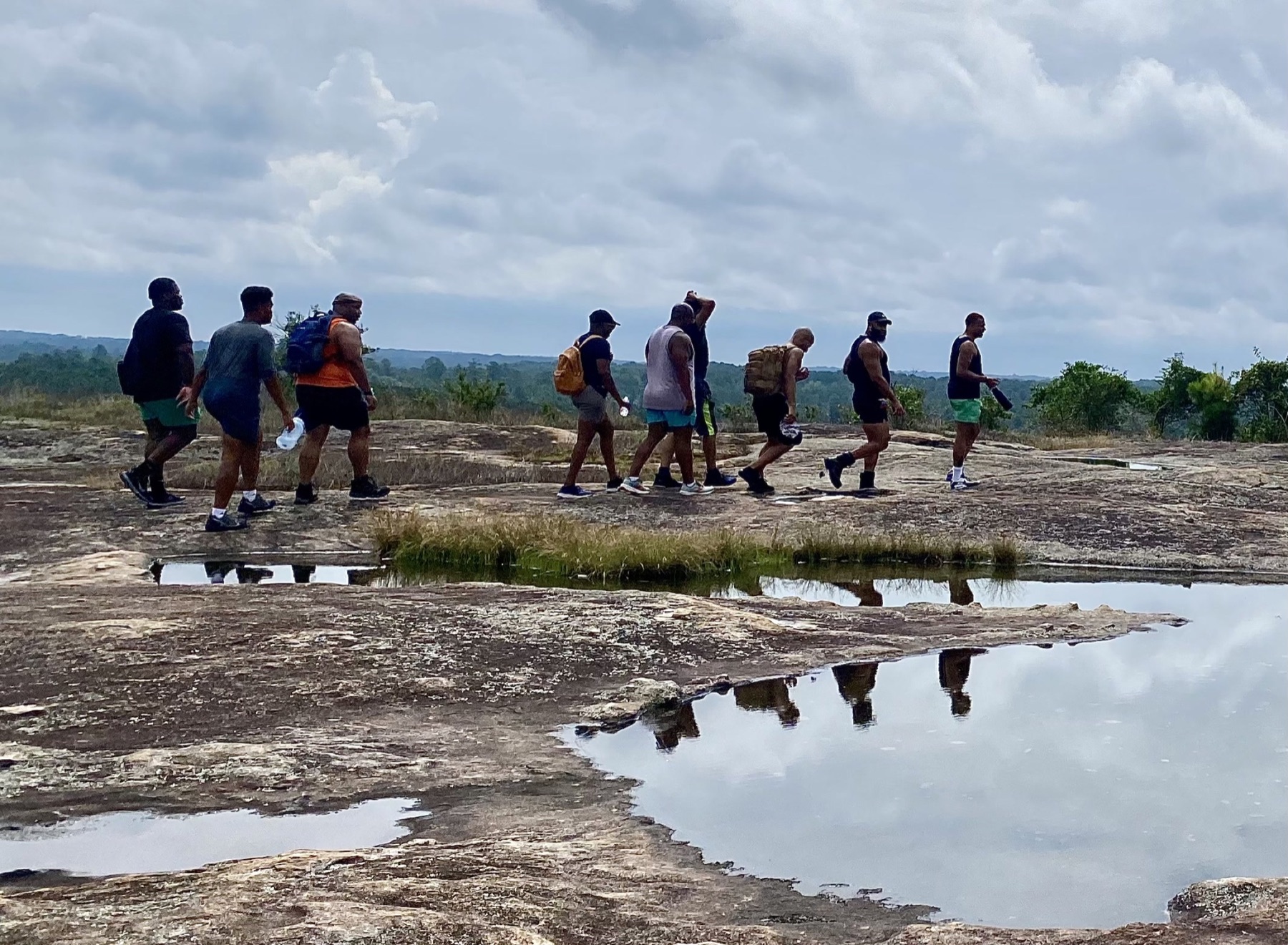 Black men cross a rain-darkened granite summit reflected in shallow pools beneath a cloudy sky.