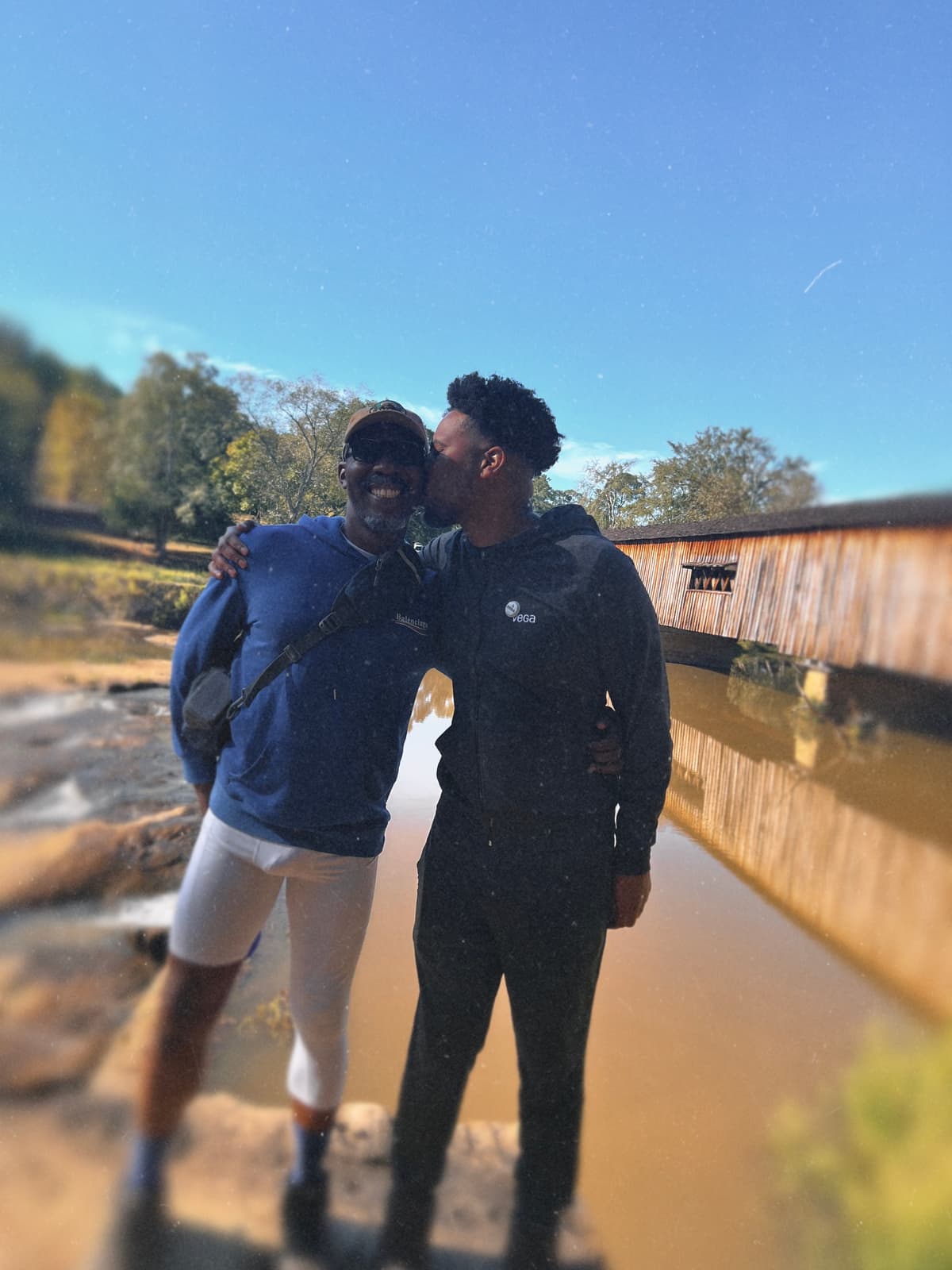 Two Black hikers stand close together beside the creek with Watson Mill’s covered bridge behind them.