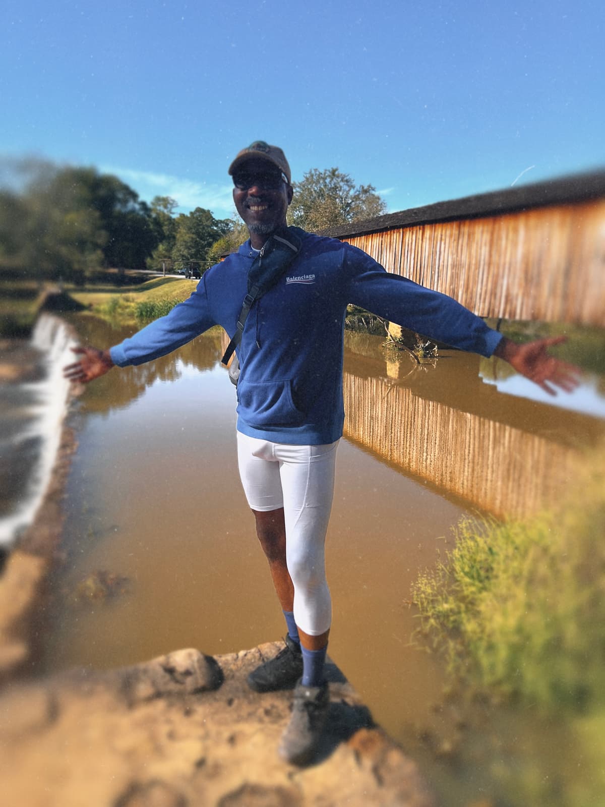 A Black hiker in a blue pullover balances with arms wide beside Watson Mill’s wooden covered bridge.