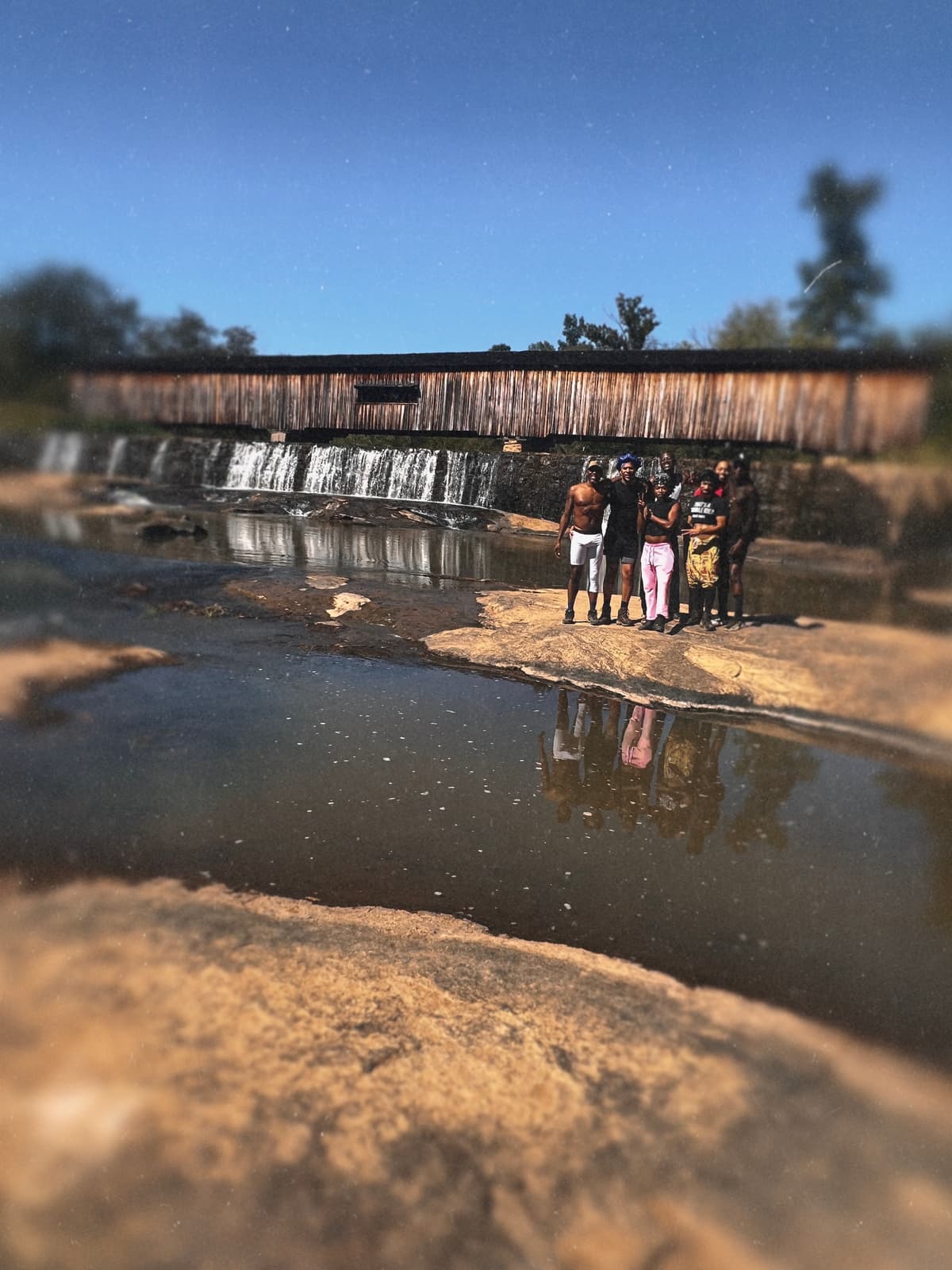 Six Black hikers stand on sunlit rock beside the waterfall with Watson Mill’s covered bridge above them.