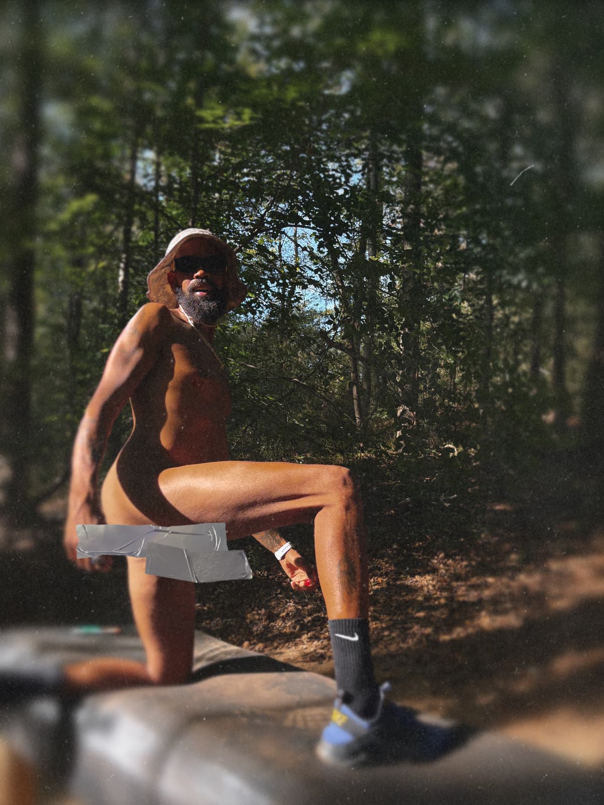 An adult Black man in clothing-optional attire sits on a fallen tree beside a shaded forest trail.