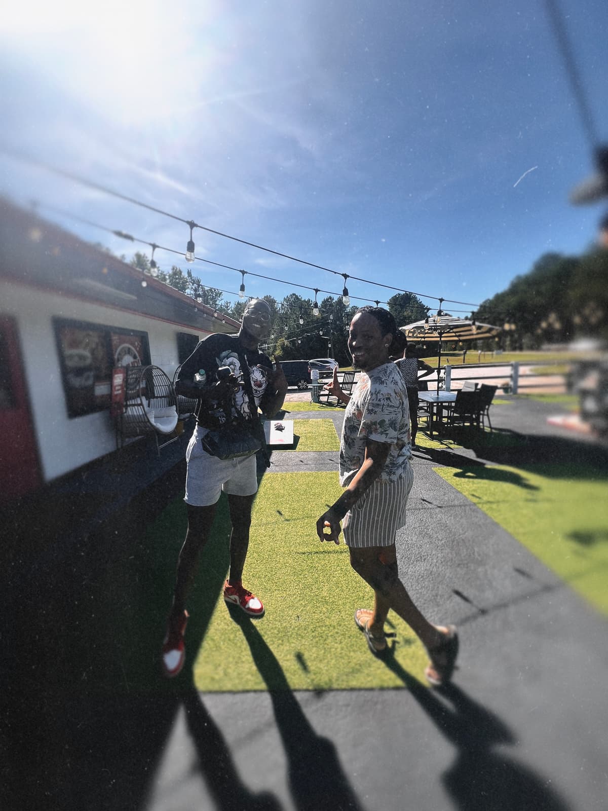 Two Black men in casual outdoor clothes talk on a sunny campground lawn beneath string lights.