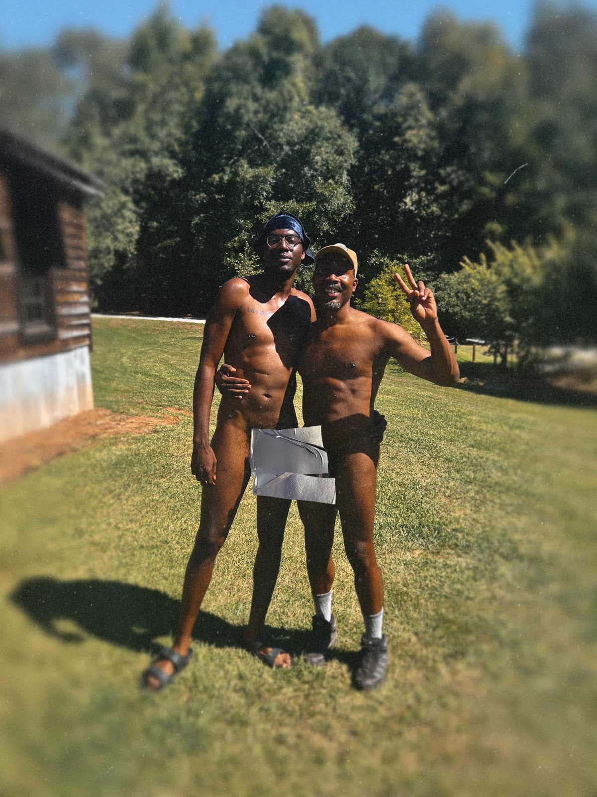 Two adult Black men in clothing-optional attire pose together on the grass beside a wooden cabin.