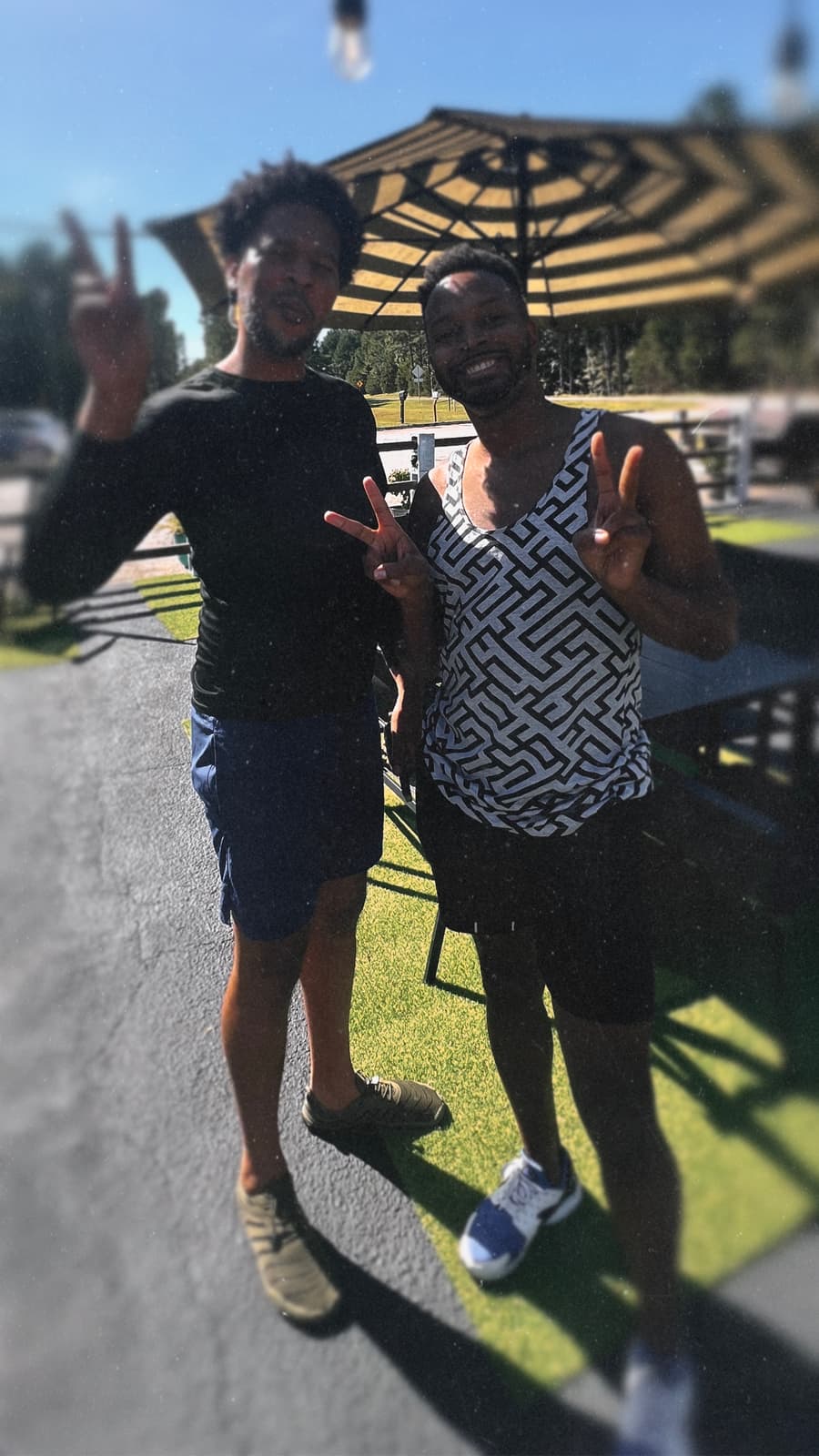 Two Black men smile together on a sunny paved path beside a campground pavilion.