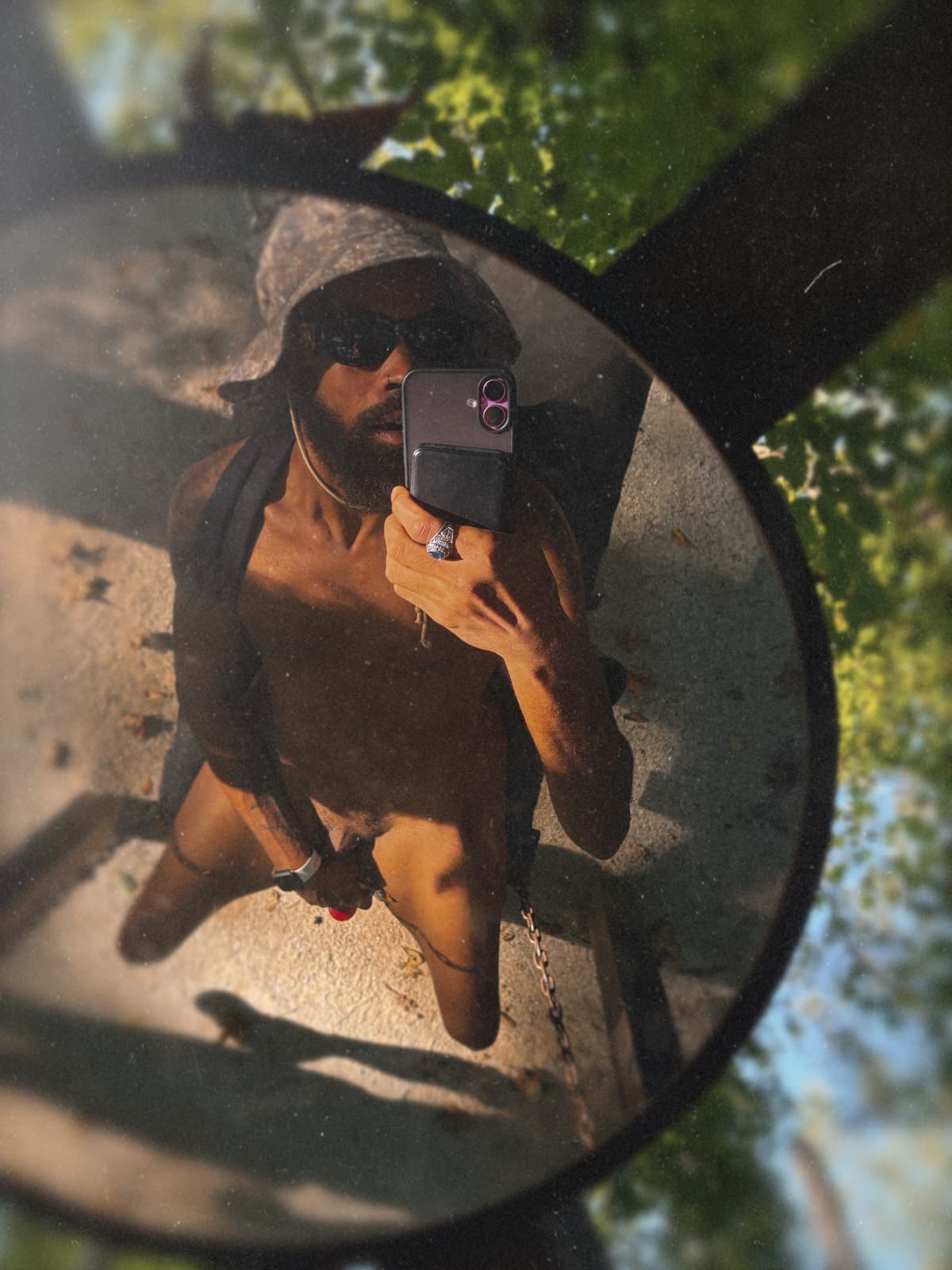 An adult Black man in black clothing takes a playful selfie reflected in a round outdoor mirror.