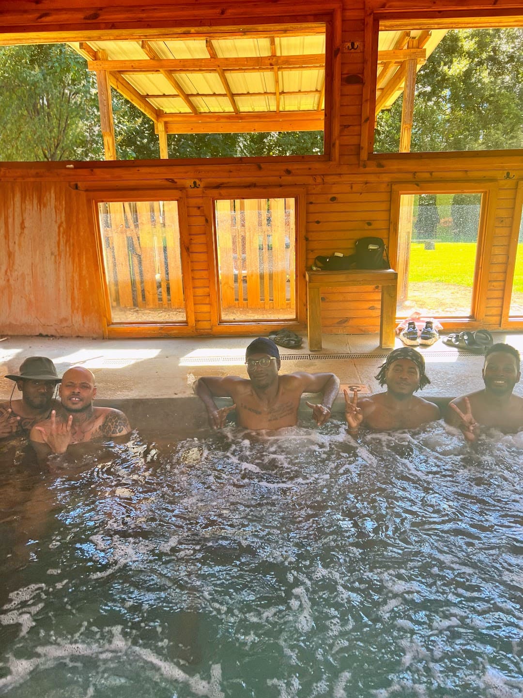 Five adult Black men smile together in a bubbling spa inside a sunlit wooden bathhouse.