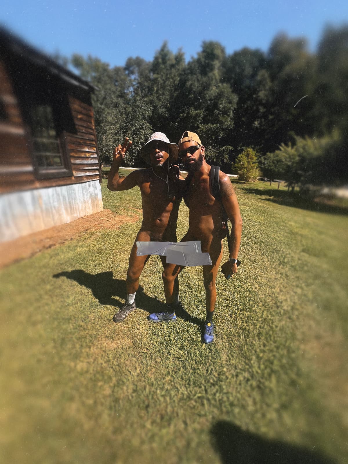 Two adult Black men in clothing-optional attire smile together on a sunny campground lawn.