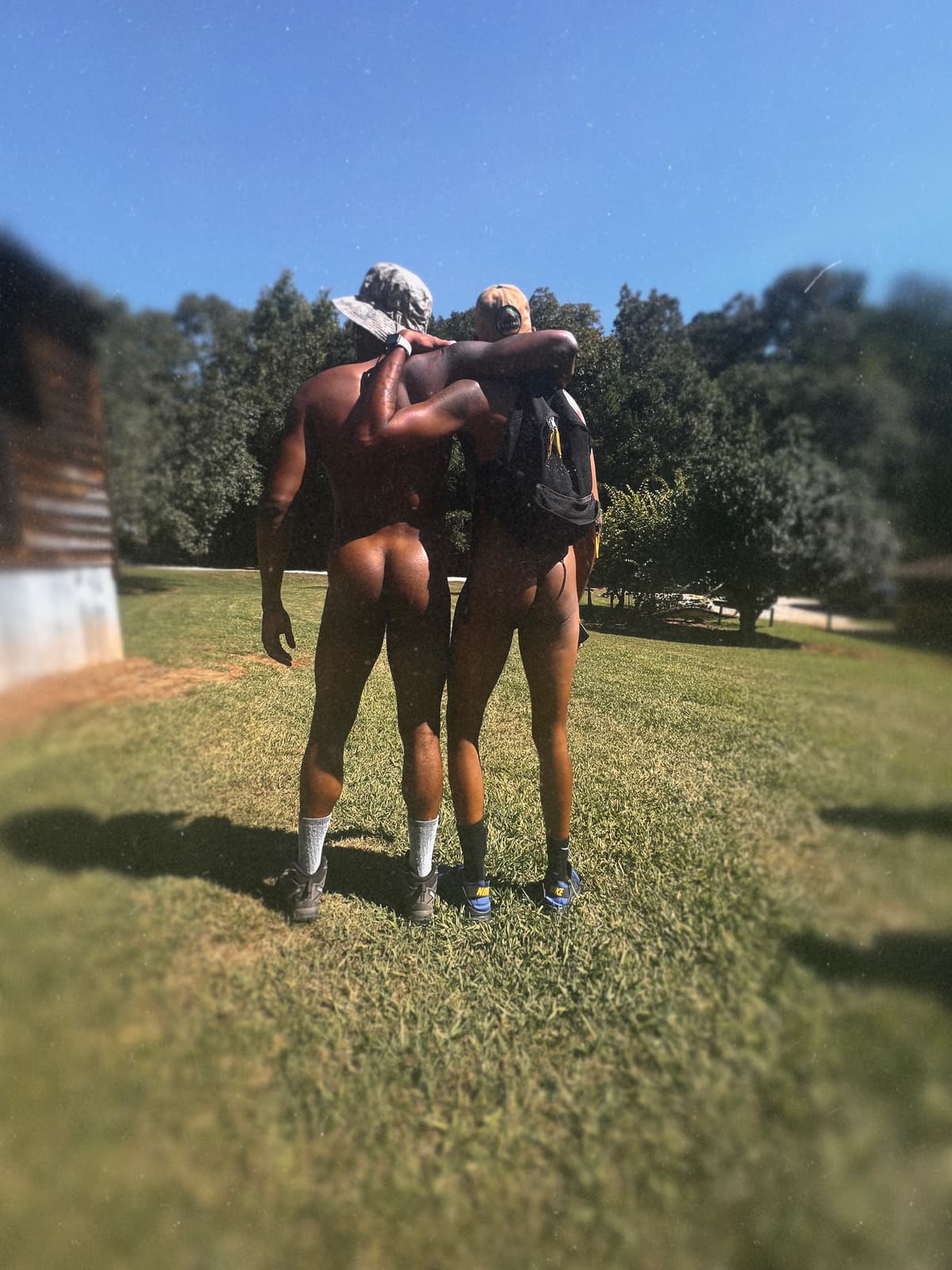 Two nude adult Black men walk away arm in arm across a grassy clothing-optional campground.