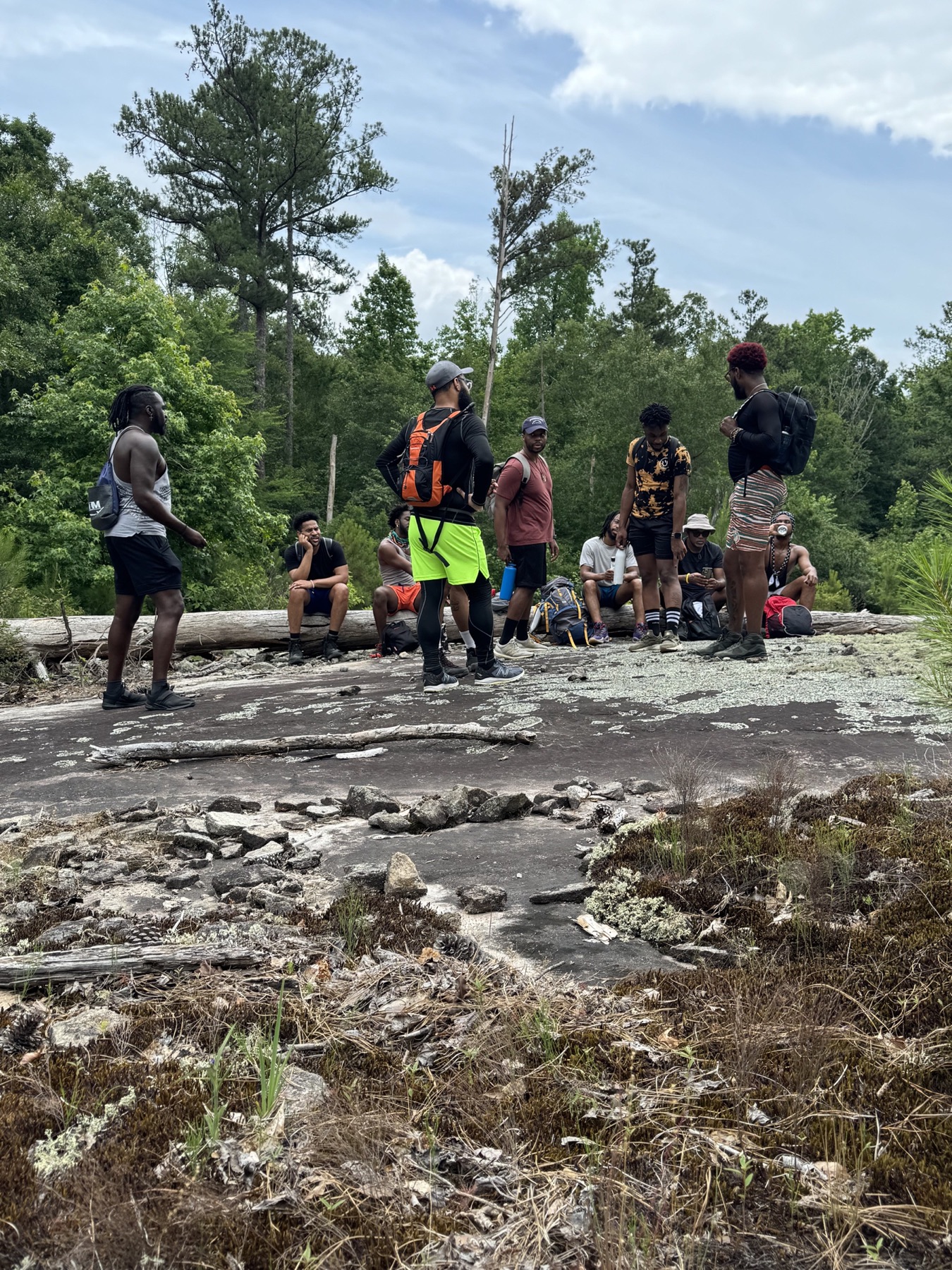 Black men gather on a rocky clearing bordered by shrubs and pine trees.
