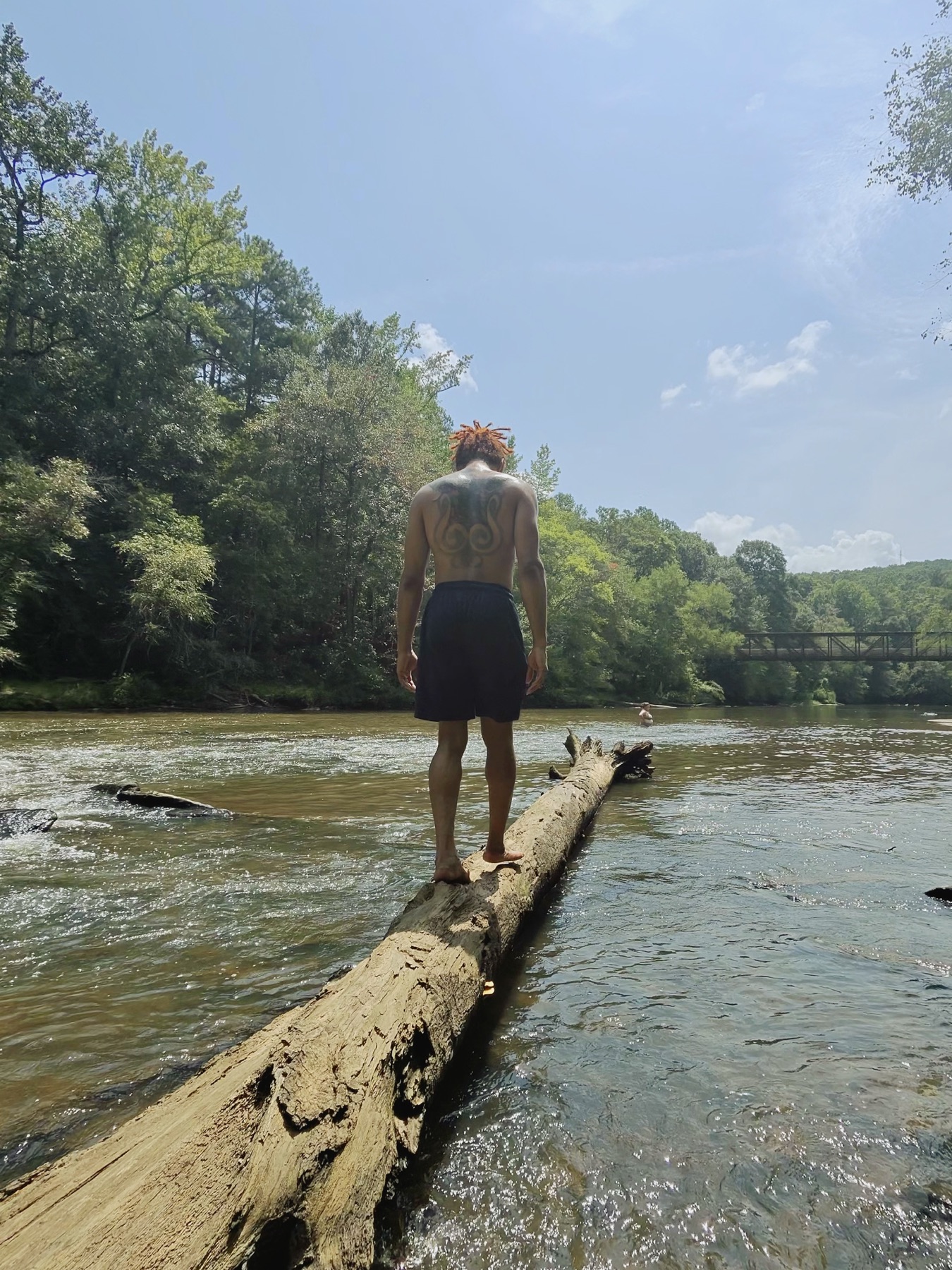 A Black man balances barefoot on a fallen log above a flowing creek with wooded banks behind him.