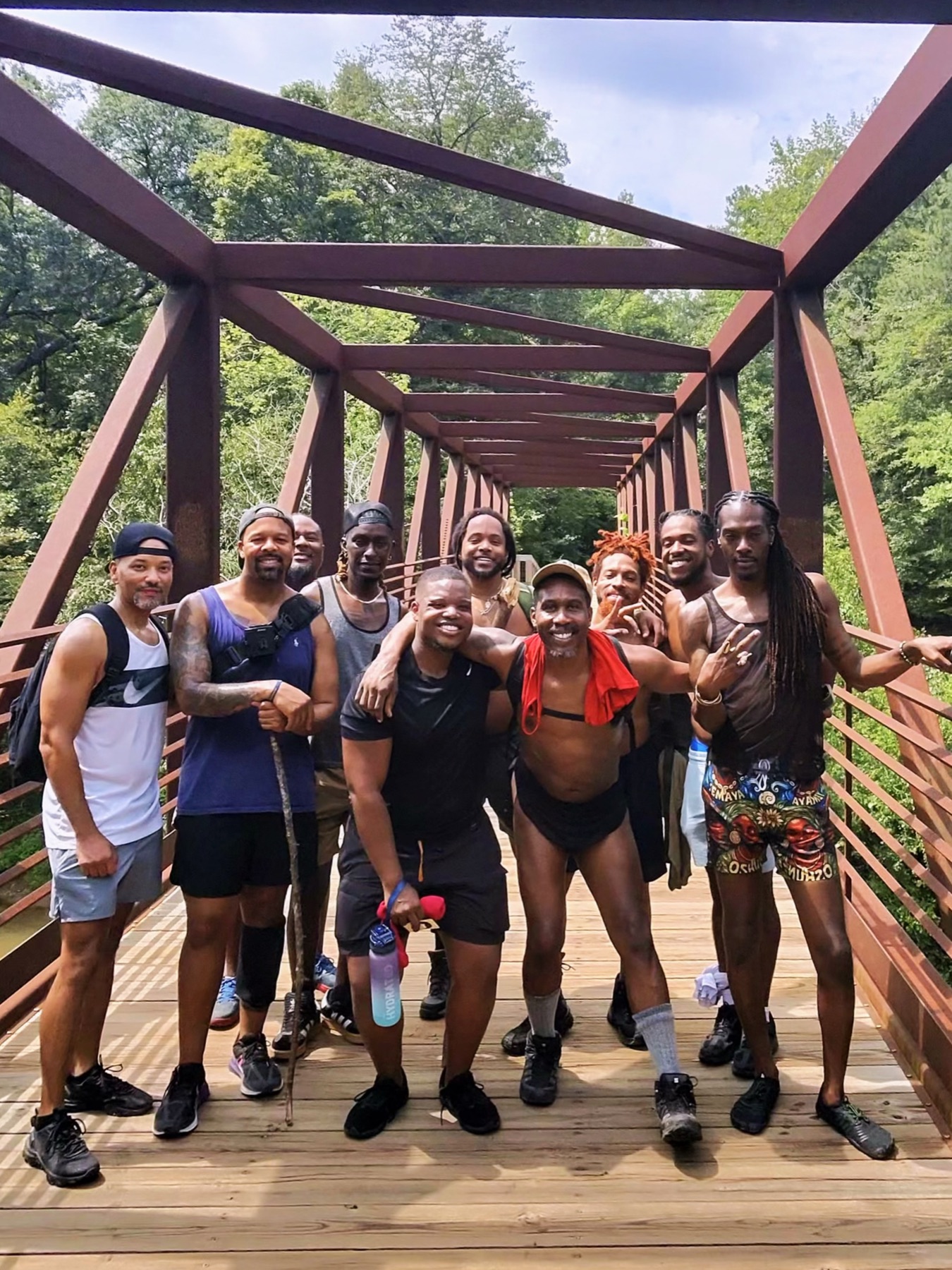 A group of Black men pose together on a rust-red steel footbridge surrounded by summer trees.