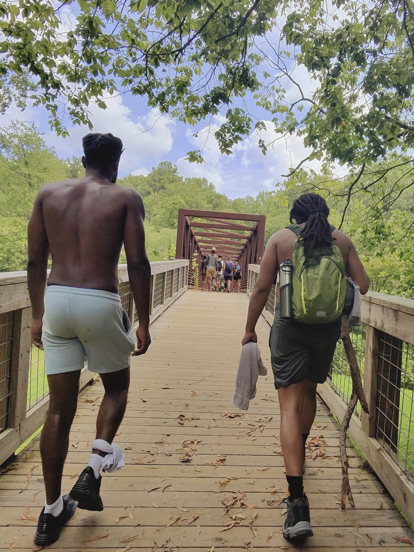 Two Black men walk toward a group crossing a red steel footbridge beneath leafy trees.