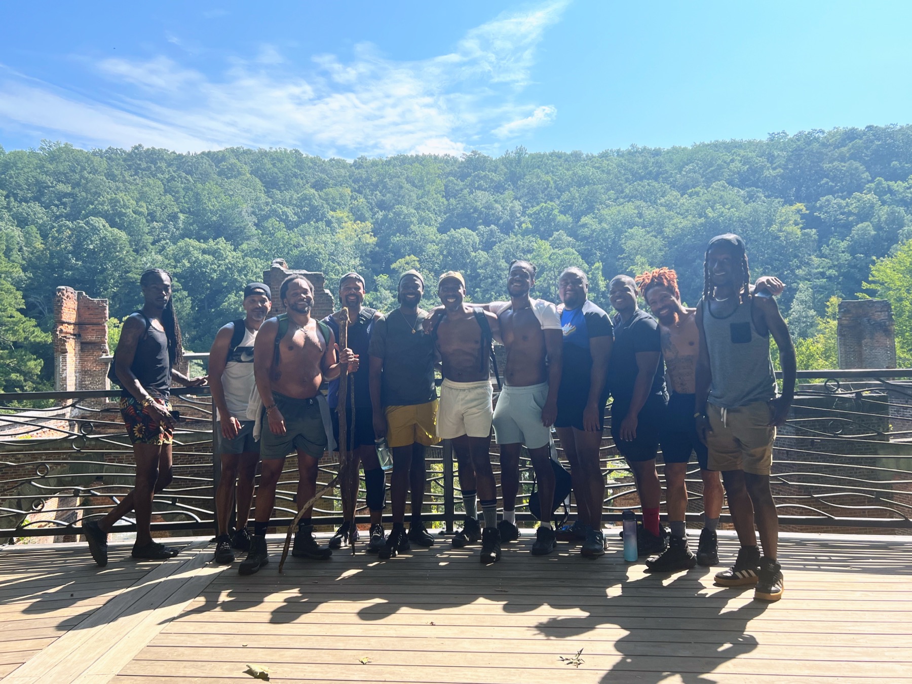 Black men stand together on an overlook above brick mill ruins and a forested valley.