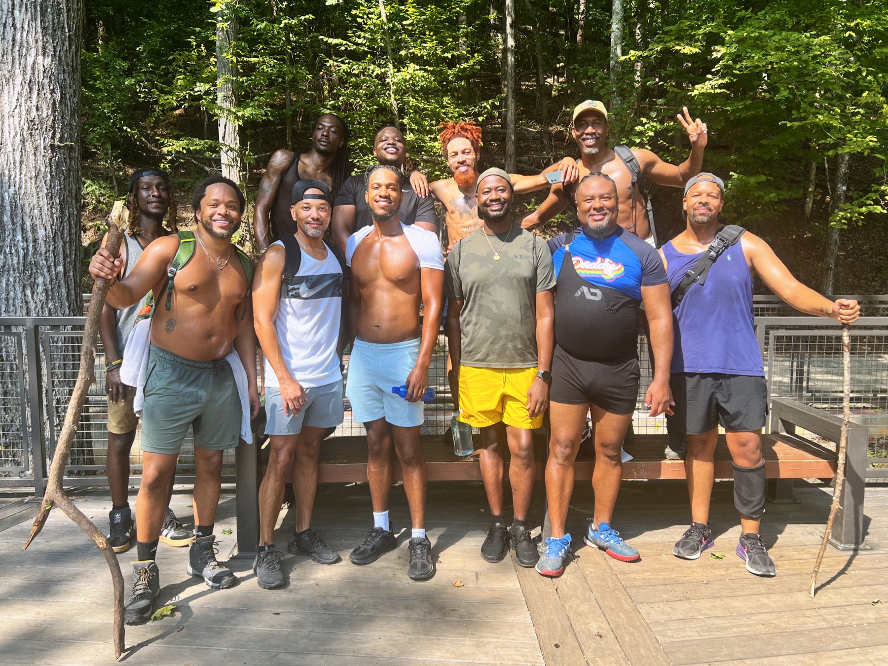 Black men smile together on a sunlit wooden deck surrounded by dense green forest.