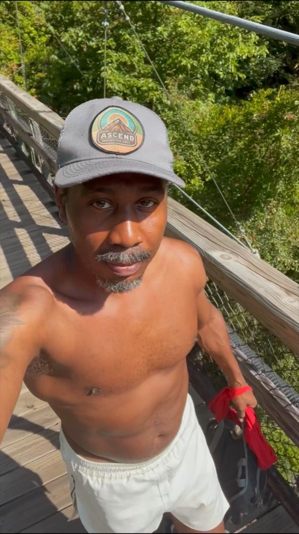 A Black hiker wearing an ASCEND cap takes a selfie on the Tallulah Gorge suspension bridge.