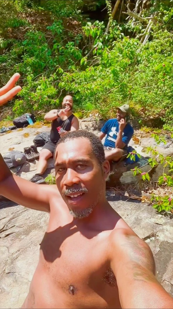 A Black hiker raises an arm for a selfie while two friends rest on sunlit rock behind him at Tallulah Gorge.