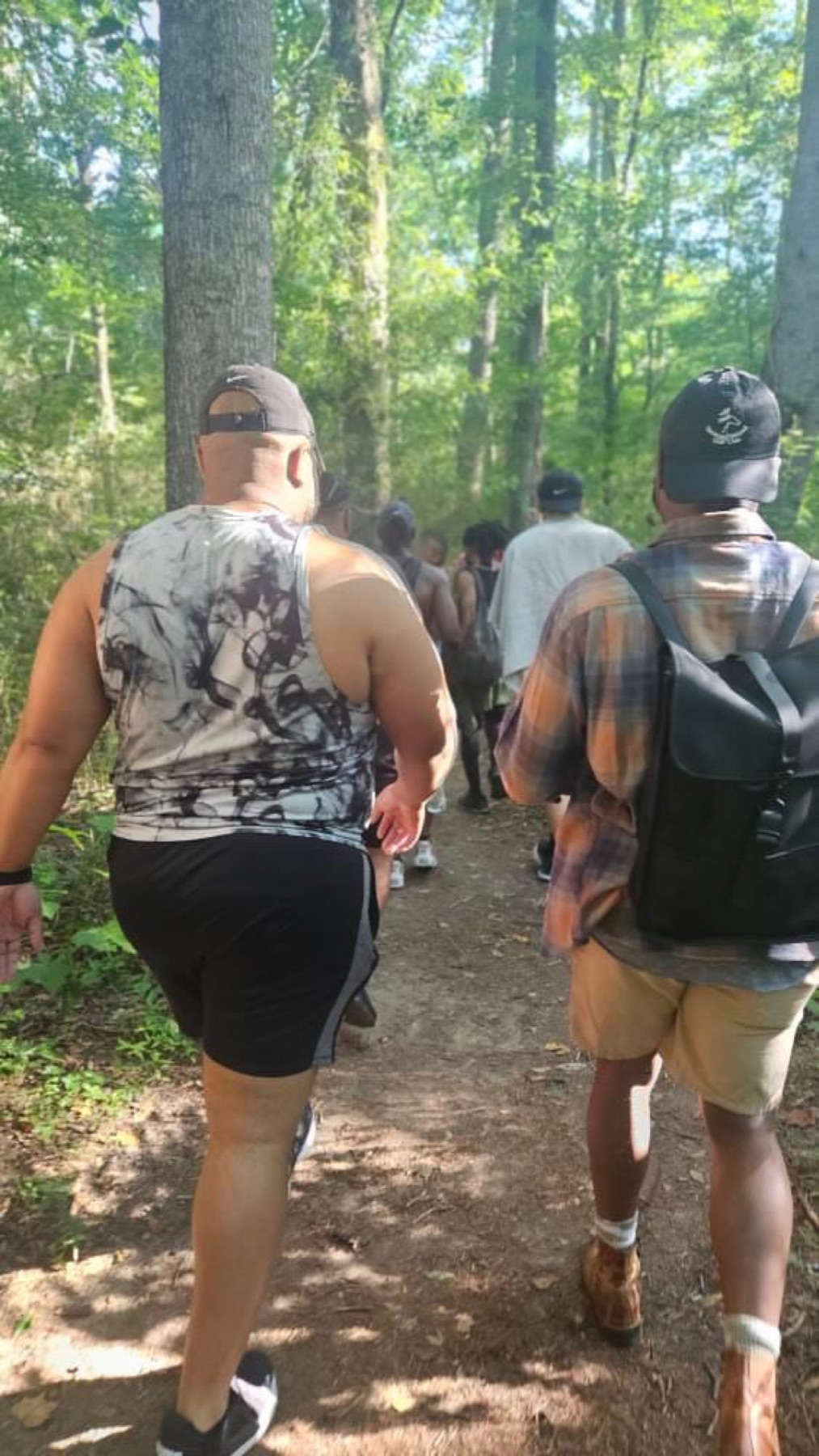 Two Black men with backpacks walk a wooded trail beneath a bright green summer canopy.