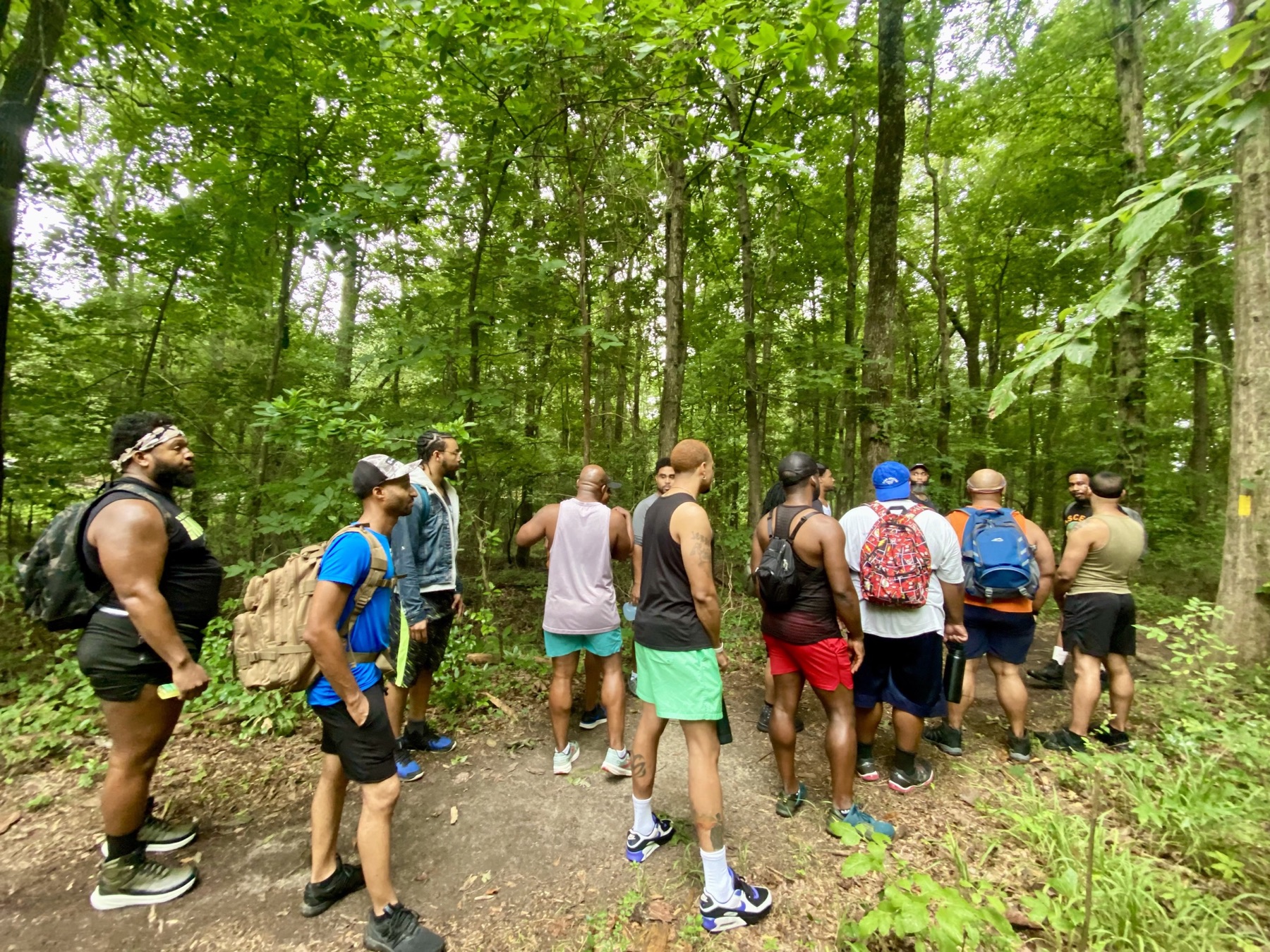 Black men with daypacks gather at the start of a green woodland trail.