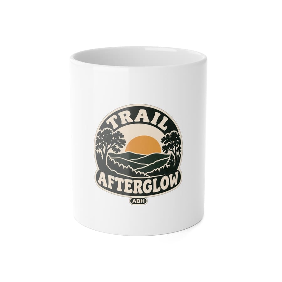 Front product mockup of a white ABH ceramic mug with a Trail Afterglow wooded-hills-and-sun badge and ABH signature.